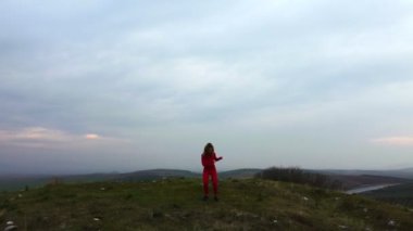 Drone footage of a 30s one woman with red tights and tops dancing on a Tumulus hill on a cloudy sky background