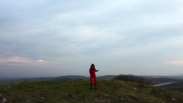 30代のドローン映像では 曇り空の背景に赤いタイツとトップスを着た女性が古墳の丘で踊っている — ストック動画