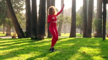 30s one woman with red tights and tops dancing at a public park on green grass with trees on the background