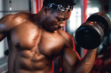 20 'li yaşlarda siyah ve kaslı bir adam spor salonunda bisepslerini kıvırıyor.