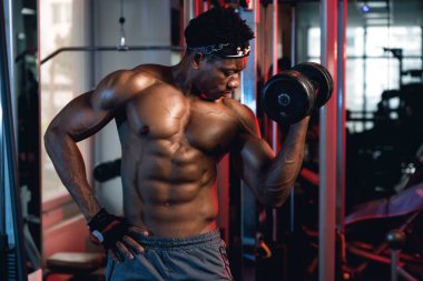 20 'li yaşlarda siyah ve kaslı bir adam spor salonunda bisepslerini kıvırıyor.