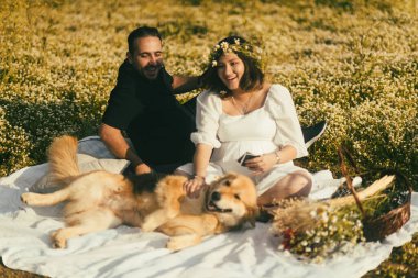 Beyaz bir piknik battaniyesinde, hamile bir kadın ve kocası bebeklerinin ultrason görüntülerini paylaşırken, dost canlısı Golden Retriever 'ları yakınlarda sevgi ararlar.