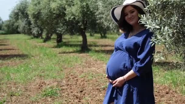 一位穿着蓝色衣服 头戴白色帽子的快乐的孕妇 在橄榄林的环境中热情地抚摸着她的腹部 与橄榄叶互动 散发着积极向上的光芒 — 图库视频影像