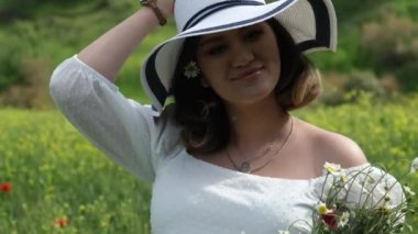Beyaz elbiseli ve şapkalı hamile bir kadının yakın plan fotoğrafı, kulağına papatya koyarken buğday tarlasında taze toplanmış papatyalar ve gelincikler tutuyor.