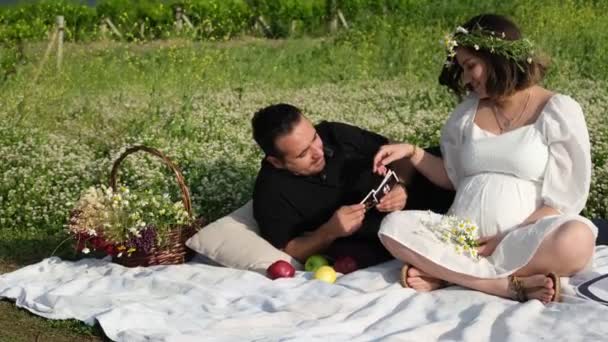 在慢镜头中 一个头戴雏菊冠的孕妇和她的黑衣伴侣坐在草地上的野餐毛毯上 抱着婴儿的超声波图像 沉浸在宁静的期待中 — 图库视频影像