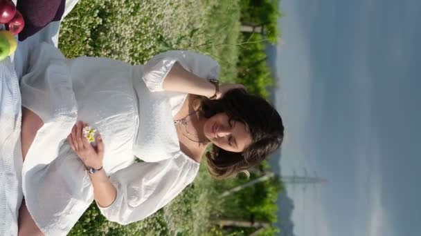 一个身穿白衣戴白帽的孕妇 坐在绿地里的白色野餐毛毯上 手里拿着一把雏菊 抱紧肚子 — 图库视频影像