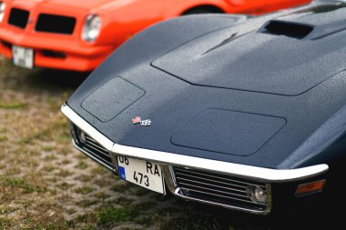 Izmir, Turkey - June 3, 2023: Close-up of the front of a black 1973 Corvette, covered in raindrops, with an out-of-focus orange Pontiac Trans Am in the background, at the IZKOD Classic Car Meet at Buca Pond. clipart