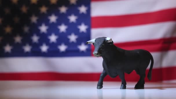 Zbliżenie Figurki Byka Symbolizujące Burzliwy Trend Rynkowy Nieostrą Amerykańską Flagą — Wideo stockowe