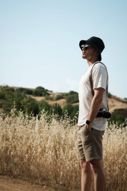 Beyaz tişört, bej şort, güneş gözlüğü ve omzuna klasik bir kamera takılmış siyah denizci şapkası giymiş yirmili yaşlarında bir adamın portresi. Yeşil tepeler ve kuru otlar arka planı oluşturur.