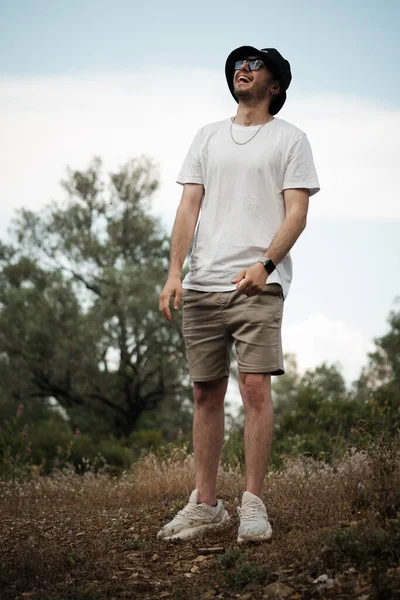 一个二十多岁的男人 穿着白色T恤 米色短裤 太阳镜和黑色水手帽 开怀大笑 他四周环绕着青山和干草的自然美 — 图库照片
