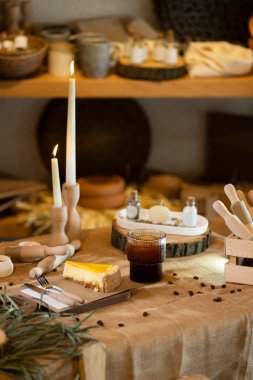 Çuval bezi masa örtüsünün üzerinde bir dilim portakallı cheesecake tahtanın üzerinde duruyor. Ahşap saplı çatal bıçak takımı ve arka planda parlayan mumlarla ortam sıcak ve çevresel bir hava yayıyor..