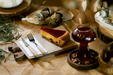 Tahta bir tahtanın üzerinde bir dilim çilekli cheesecake 'in bir fincan kış çayının yanında sergilendiği sıcak bir sahne. Mum ışığı altında bir çuval bezi koyun, bu ortam rahatlık ve hoşgörüyü temsil eder..