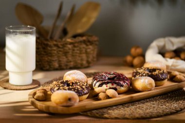 Tahta bir tepside üç çörek ve ısırık büyüklüğünde bisküvilerle çikolata dolu kurabiyeler var. Beyaz kumaş arka planda bir bardak süt ve bir sepet ahşap mutfak aletinin yanında ceviz bulundurur..