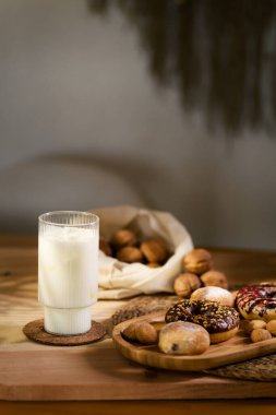 Tahta bir tepside üç çörek ve ısırık büyüklüğünde bisküvilerle çikolata dolu kurabiyeler var. Beyaz kumaş arka planda bir bardak sütün yanında ceviz saklar..