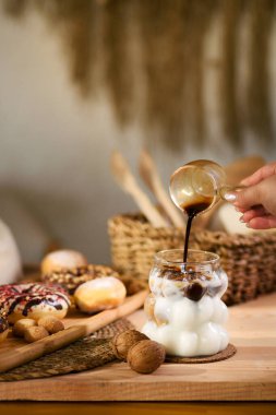 Bir kadının elinin cam bir kahve demliğinden buzlu sütle dolu bir bardağa espresso döktüğü anı yakalayan bir fotoğraf..