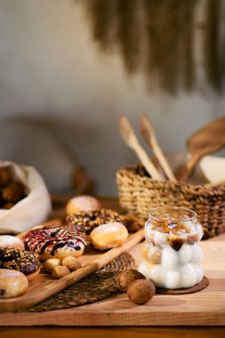 Tahta bir tabakta donutlar, çikolata dolu kurabiyeler ve ısırık büyüklüğünde kurabiyeler var. Arkada, cevizler beyaz bir bezin üzerinde duruyor, sepetteki ahşap çatal bıçaklarla. Ekspreso ile buzlu süt sahneyi tamamlıyor.