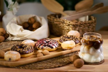 Tahta bir tabakta donutlar, çikolata dolu kurabiyeler ve ısırık büyüklüğünde kurabiyeler var. Arkada, cevizler beyaz bir bezin üzerinde duruyor, sepetteki ahşap çatal bıçaklarla. Ekspreso ile buzlu süt sahneyi tamamlıyor.