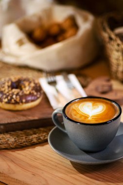 Tahta bir tahtanın üzerinde, gri bardaktaki lattenin yanında bir çörek. Ahşap çatal bıçaklı beyaz peçete ve arka planda beyaz kumaşla kabuklu fındık..