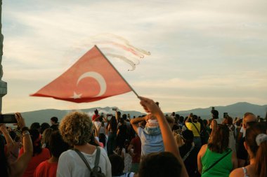 İzmir, Türkiye, 9 Eylül 2023: Akşam güneşi altın ışığını saçarken, Türk Yıldızları bir hava gösterisi sergiliyor. Türk bayrakları taşıyan büyülenmiş izleyiciler, İzmi 'nin yıldönümü münasebetiyle ikonik Cumhuriyet Ağacı anıtının etrafında toplandılar.