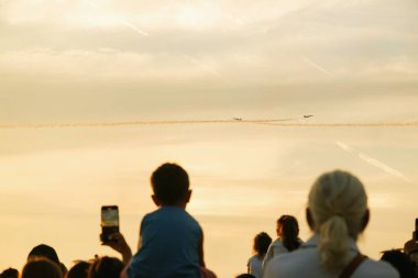 İzmir, Türkiye, 9 Eylül 2023: Akşam güneşi altın ışığını saçarken, Türk Yıldızları bir hava gösterisi sergiliyor. Türk bayrakları taşıyan büyülenmiş izleyiciler, İzmi 'nin yıldönümü münasebetiyle ikonik Cumhuriyet Ağacı anıtının etrafında toplandılar.