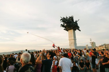 İzmir, Türkiye, 9 Eylül 2023: Akşam güneşi altın ışığını saçarken, Türk Yıldızları bir hava gösterisi sergiliyor. Türk bayrakları taşıyan büyülenmiş izleyiciler, İzmi 'nin yıldönümü münasebetiyle ikonik Cumhuriyet Ağacı anıtının etrafında toplandılar.