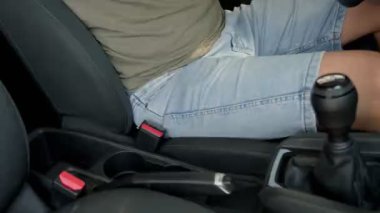 Yolcunun perspektifinden alınan görüntülerde kot şortlu bir erkek sürücü arabanın içinde el frenini çekiyor ve rutin güvenlik önlemlerini vurguluyor..