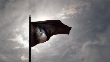 Türk bayrağının siluet olarak dalgalandığı yakın çekim ve yavaş çekim bir video, bulutlu bir gökyüzüne karşı çekilmiş, güneş içeri bakıyor, bir dramatı yakalıyor