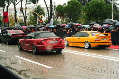 Tirana, Albania - November 28, 2023: On Avenue of Martyrs of the Nation, citizens watch a modified car parade on a rainy Independence Day, featuring a red and yellow BMW 3 E46 and a black colored dodge challenger labeled Cair Tuning Team clipart