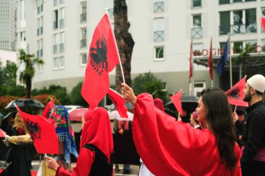 Tiran, Arnavutluk - 28 Kasım 2023: Ulusun Şehitleri Bulvarı 'nda, geleneksel giyinmiş öğrenci ve insanlar, arka planda şemsiye ve bayraklar tutan izleyicilerle yağmurlu bir Bağımsızlık Günü' nde Arnavut bayraklarıyla coşkuyla yürüyorlar.