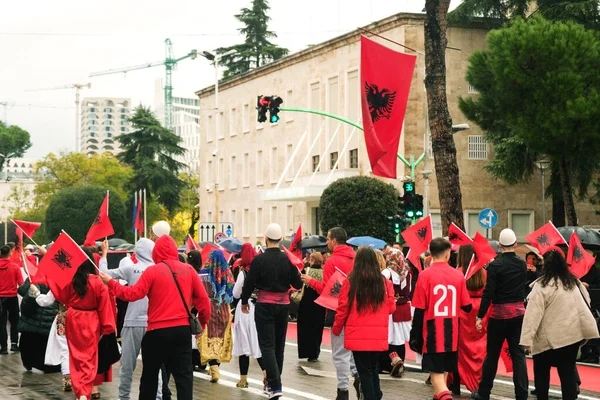 Tiran, Arnavutluk - 28 Kasım 2023: Ulusun Şehitleri Bulvarı 'nda, geleneksel giyinmiş öğrenci ve insanlar, arka planda şemsiye ve bayraklar tutan izleyicilerle yağmurlu bir Bağımsızlık Günü' nde Arnavut bayraklarıyla coşkuyla yürüyorlar.