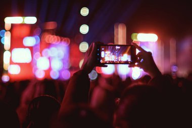 Bir kişi gece Skanderberg Meydanı 'nda arka planda Bokeh sahne ışıkları olan bir akıllı telefonla konser kaydediyor.