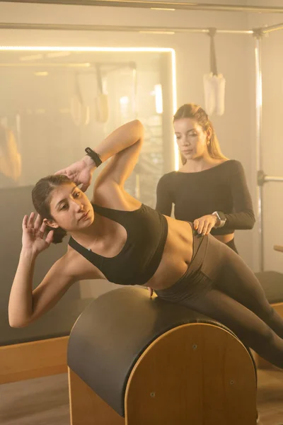 A young woman performs core exercises on a Reformer Pilates machine in a gym, guided by a trainer in the Reformer room