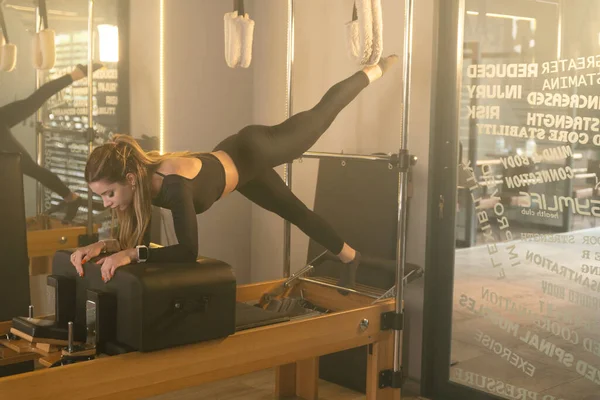 A young woman performs a leg lift core exercise on a Reformer Pilates machine in a gym, targeting core strength and flexibility