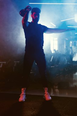 Bir adam spor salonunda kettlebell kapma gösterisi yapıyor. Sanatsal mavi ve kırmızı ışıklı ve ince sisli bir atmosferi var.