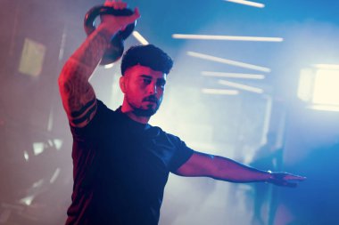 Bir adam spor salonunda kettlebell kapma gösterisi yapıyor. Sanatsal mavi ve kırmızı ışıklı ve ince sisli bir atmosferi var.