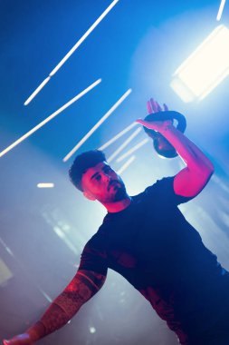 Bir adam spor salonunda kettlebell kapma gösterisi yapıyor. Sanatsal mavi ve kırmızı ışıklı ve ince sisli bir atmosferi var.