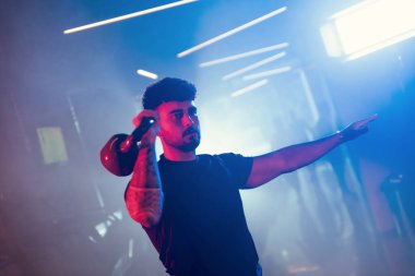 Bir adam spor salonunda kettlebell kapma gösterisi yapıyor. Sanatsal mavi ve kırmızı ışıklı ve ince sisli bir atmosferi var.