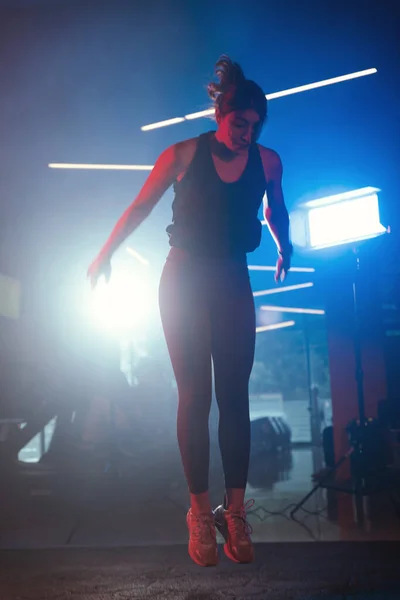 Capturing the peak of a burpee jump, a woman trains under vibrant blue and red gym lights with a misty ambiance