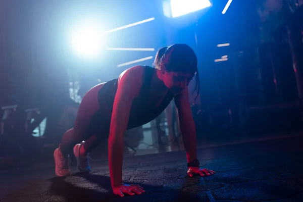 Capturing the peak of a burpee jump, a woman trains under vibrant blue and red gym lights with a misty ambiance