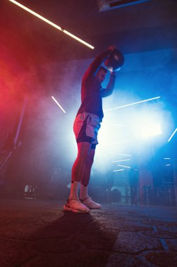 Erkek bir sporcunun kırmızı ve mavi ışıkların sisli parlaklığıyla çevrili bir ilaç topunu yere çarptığı yoğun bir çalışma anı.