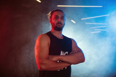 Spor salonunda kollarını kavuşturan erkek bir sporcunun portresi. Mavi ve kırmızı ışıklarla işaretlenmiş, sisli, arka plan aydınlatmalı.