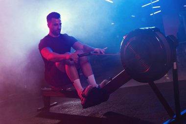 Erkek bir sporcu kürek çekme makinesine kürek çeker. Mor mavi ve kırmızı ışıklarla aydınlanır. Hafif sisle örtülüdür.