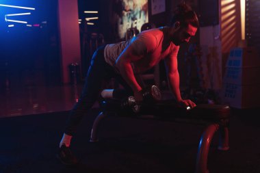 Odaklanmış erkek sporcu, havada ince bir sis ile mavi ve kırmızı ışıkların dinamik bir oyunu arasında halterli sıra egzersizi yapıyor..