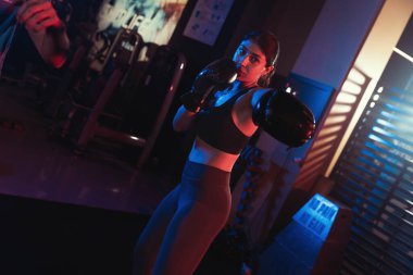 Kendini adamış bir bayan sporcu, sislerle dolu bir spor salonunda, dramatik mavi ve kırmızı ışıklar altında yumruklarını çalışıyor.