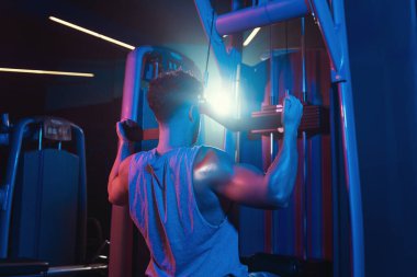 Bir adam formunu, spor salonunun mistik mavi ve kırmızı renkleri arasında yakalanan odaklanma ve güç kaynaşma makinesinde mükemmelleştirir.