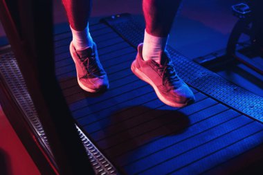 Puslu bir ortamda dramatik mavi ve kırmızı spor salonu ışıklarıyla vurgulanan koşu bandında spor ayakkabılarına yaklaş.