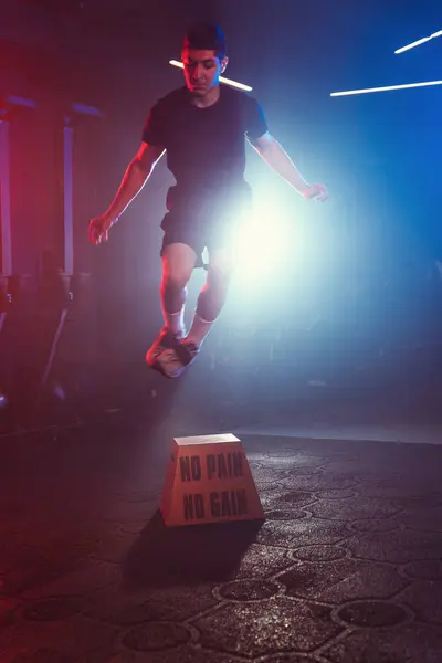 An athlete executes a box jump, soaring above \'No Pain No Gain,\' amidst a gym aglow with blue and red lights and soft mist
