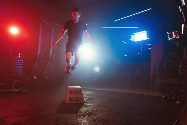 An athlete executes a box jump, soaring above \'No Pain No Gain,\' amidst a gym aglow with blue and red lights and soft mist