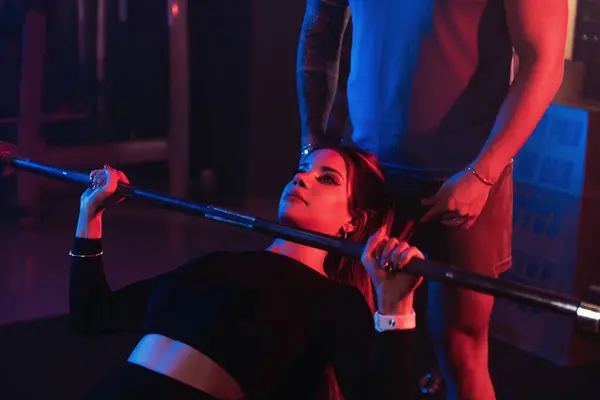 A dedicated female athlete engages in an incline bench press, guided by her trainer amidst the dramatic gym setting highlighted by blue and red lights