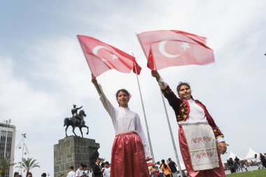 İzmir, Türkiye - 23 Nisan 2024 İki neşeli kız Çocuk Günü kutlamalarında geleneksel gösteri elbiseleriyle Türk bayrağını sallıyor, arka planda at sırtında Atatürk heykeli